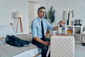 Handsome mature man in shirt and tie leaning at the suitcase while sitting on bed at home photo