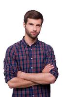 casualmente guapo. retrato de un apuesto joven con camisa informal que mantiene los brazos cruzados y mira la cámara mientras está aislado en blanco foto