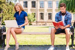 Relaxing separately. Handsome young man sitting on the bench and reading book while beautiful woman sitting near him and using computer photo