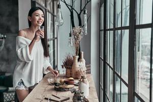 Day starts with healthy breakfast. Attractive young woman drinking water with lemon and smiling while standing near the window at home photo