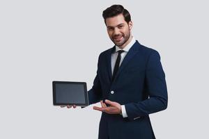 Just take a look. Good looking young man in full suit pointing at digital tablet and looking at camera with smile while standing against grey background photo