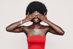 Beautiful young African woman covering eyes with hands and smiling while standing against gray background photo