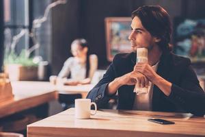 Lost in thoughts. Handsome young man holding newspaper and looking thoughtful while sitting in cafe photo