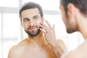 Skin care. Handsome young shirtless man applying cream at his face and looking at himself with smile while standing in front of the mirror photo