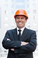 Confident contractor. Cheerful mature man in formalwear and hardhat keeping arms crossed and smiling while standing outdoors photo