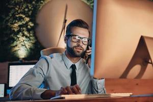 Confident young man working on computer and talking on mobile phone while staying late in the office photo
