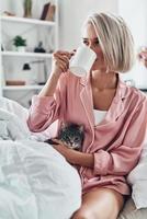 Morning coffee. Attractive young woman drinking coffee and holding her cat while sitting in bed at home photo