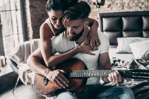 Playing by heart. Handsome young bearded man sitting in bed and playing guitar while attractive woman embracing him photo