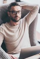Handsome and thoughtful. Handsome man using his laptop and looking pensive while sitting in chair photo