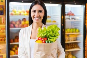 feliz de encontrar todo lo que quiero. hermosa joven sosteniendo una bolsa de compras con frutas y sonriendo mientras está de pie en la tienda de comestibles cerca del refrigerador foto