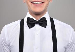 Close-up on nerd. Cropped image of young nerd man in bow tie and suspenders smiling while standing against grey background photo