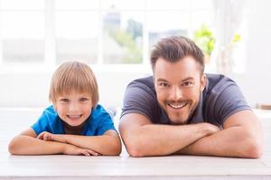 el hogar es donde está el padre. feliz padre e hijo inclinando sus rostros en las manos y sonriendo mientras yacen en el suelo en casa foto