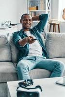 Just relaxing. Handsome young African man holding television set and smiling while sitting indoors photo