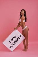 Smile to yourself every day.  Full length of attractive young smiling woman holding a poster and looking at camera while standing against pink background photo