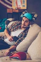 Young handsome man in beer hat holding joystick and looking at camera while sitting on sofa in messy room after party photo