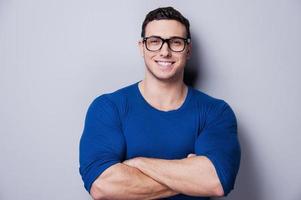 confiado y creativo. un joven alegre con los brazos cruzados y sonriendo a la cámara mientras se enfrenta a un fondo gris. foto