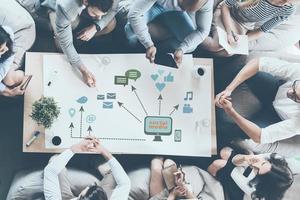 Discussing new strategy. Top view of business people discussing something while sitting around the desk together and pointing large paper with conceptual business icons drawn on it photo