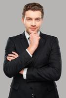 Thinking about business. Thoughtful young man in formalwear looking at camera while holding hand on chin and standing against grey background photo