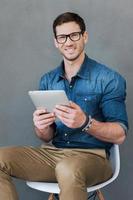 Wireless freedom. Happy young man holding digital tablet and looking at camera while sitting against grey background photo