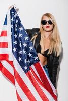 estrella americana hermosa mujer joven en sujetador negro y chaqueta de cuero sosteniendo la bandera americana y mirando a la cámara mientras está de pie contra el fondo blanco foto