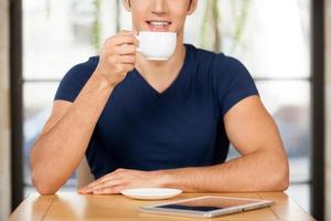 Drinking coffee at restaurant. Cropped image of cheerful young man drinking coffee at the restaurant and smiling while digital tablet laying on the table photo