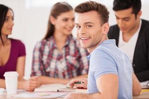 Feeling confident about his final exam. Four cheerful students sitting together at the desk and studying while one man looking over shoulder and smiling photo