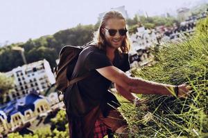 Real explorer. Handsome young man in casual clothing looking at camera and smiling while standing on the hill with buildings in the background photo