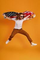 Confident young man in casual clothing making a face and looking away while holding American flag against yellow background photo