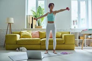 Confident young woman in sports clothing exercising with dumbbells at home photo