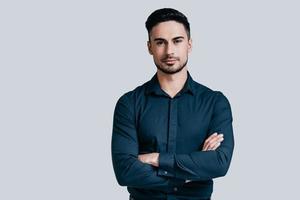 Confident and handsome. Handsome young man keeping arms crossed and looking at camera while standing against grey background photo