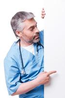 Surgeon pointing copy space. Confident mature surgeon pointing copy space while standing isolated on white photo