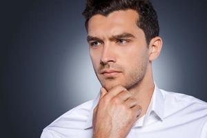 Thinking about solutions. Handsome young man in white shirt looking away and holding hand on chin while standing against grey background photo