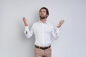 un joven apuesto con camisa blanca mirando a la cámara y gesticulando mientras se enfrenta a un fondo gris foto