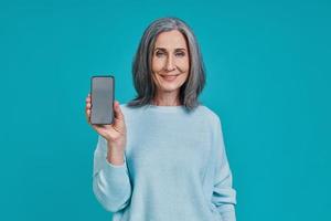 hermosa mujer madura mostrando un teléfono inteligente y sonriendo mientras está de pie contra el fondo azul foto