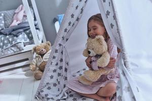 una niña encantadora abrazando a su oso de juguete y manteniendo los ojos cerrados mientras se sienta en el suelo en una tienda foto