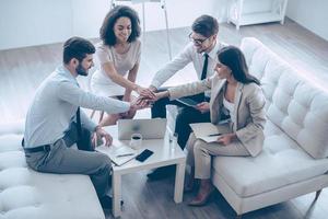 Ready for new achievements. Top view of group of four young people holding hands and showing their unity with smile while sitting on the couch at office photo