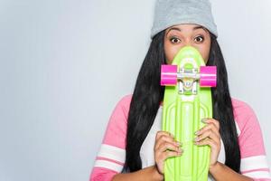 Hi there Beautiful young African woman in funky clothes hiding behind her colorful skateboard while standing against grey background photo