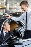 Let me show you all the features. Confident mature man ion formalwear sitting at the front seat of the car and talking to the car salesman standing near him photo