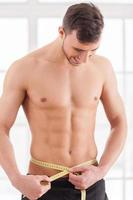 Keeping body in perfect shape. Young muscular man measuring his waist with measuring tape and smiling photo