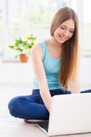 pasar un buen rato en casa. alegre adolescente usando computadora y sonriendo mientras se sienta en el suelo en su apartamento foto