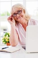 sentirse joven y activo. alegre anciana ajustándose las gafas y sonriendo mientras trabaja en una laptop foto