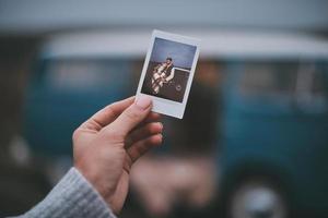 forma perfecta de recordar los mejores momentos. primer plano de una mujer joven sosteniendo una foto de una pareja joven mientras está de pie al aire libre cerca de la mini furgoneta de estilo retro azul