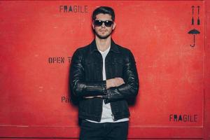 Trendy handsome. Handsome young man in sunglasses keeping arms crossed and looking at camera while standing against red background photo