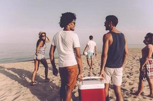 listo para la fiesta en la playa. grupo de jóvenes alegres caminando por la playa hacia el mar mientras dos hombres llevan un refrigerador de plástico foto