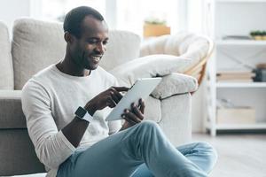navegando en la red. apuesto joven africano tocando su tableta digital y sonriendo mientras se sienta en la alfombra en casa