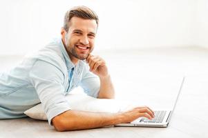 Spending time at home. Cheerful young man working on laptop and looking at camera while lying on the floor at his apartment photo