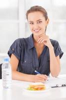 Busy beauty. Beautiful young woman writing something in her note pad and smiling while sandwich and a bottle of water laying on the table photo