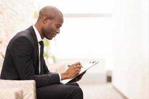 Making notes. Side view of cheerful young African businessman writing something in his note pad and smiling while sitting on the chair photo