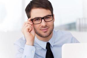 How may I help you Cheerful young man in shirt and tie looking out of the laptop and adjusting his glasses while sitting at his working place photo
