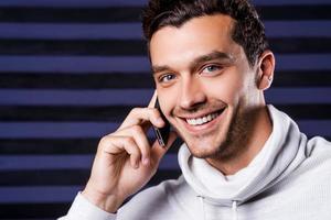 hombre guapo en el teléfono. un joven apuesto con suéter blanco hablando por teléfono móvil y sonriendo mientras se enfrenta a un fondo rayado foto